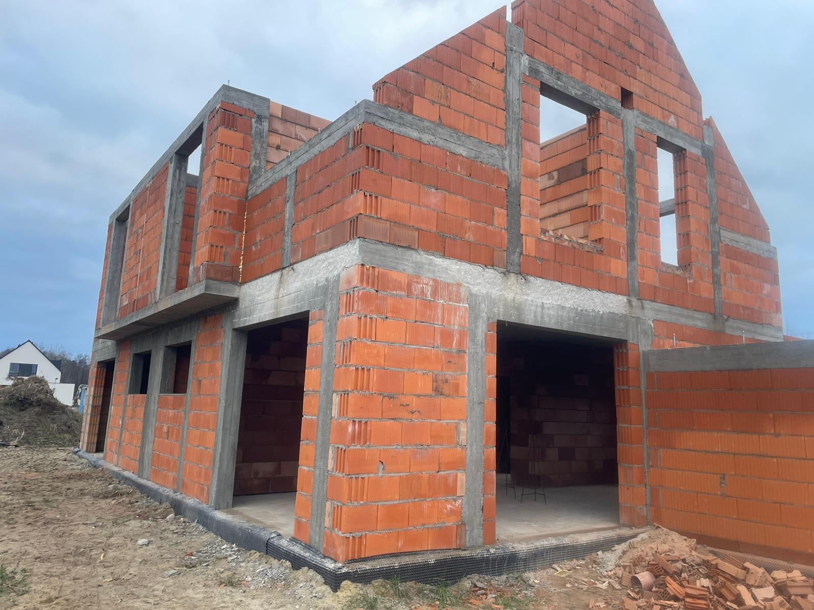 Surowy stan domu z czerwonej cegły w Otwocku, z widocznymi otworami okiennymi i betonowymi nadprożami, na tle pochmurnego nieba. Widoczny fundament i teren budowy.