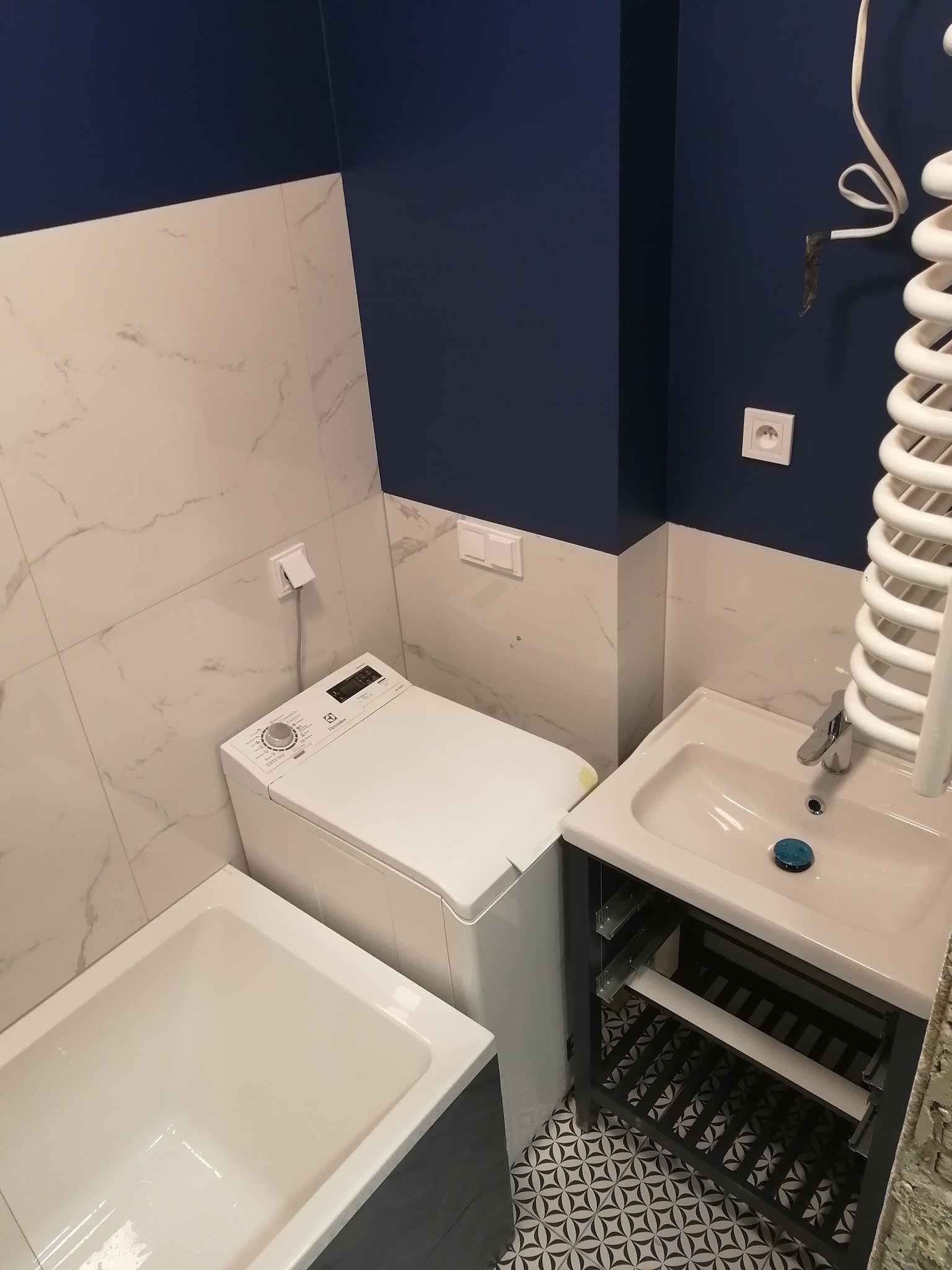 Wykończona łazienka w odcieniach bieli i granatu: biała wanna, pralka Electrolux, umywalka z szafką na tle płytek imitujących marmur i granatowej ściany z wystającym kablem.