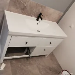 Biała szafka pod umywalkę z czarną armaturą na tle płytek imitujących marmur, widoczne elementy montażowe szuflady.