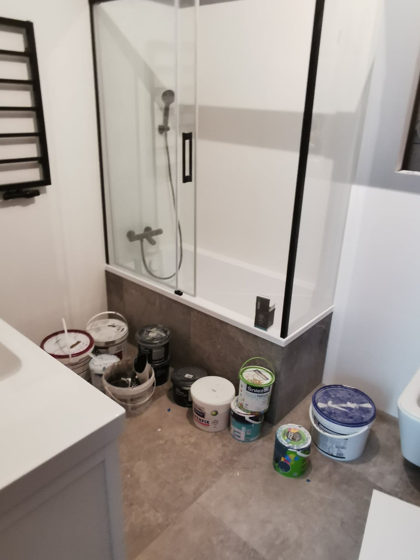 Łazienka w trakcie prac w Strzepczu: widoczna wanna z czarną ramą kabiny prysznicowej, grzejnik drabinkowy oraz liczne puszki z farbami i zaprawami na podłodze.