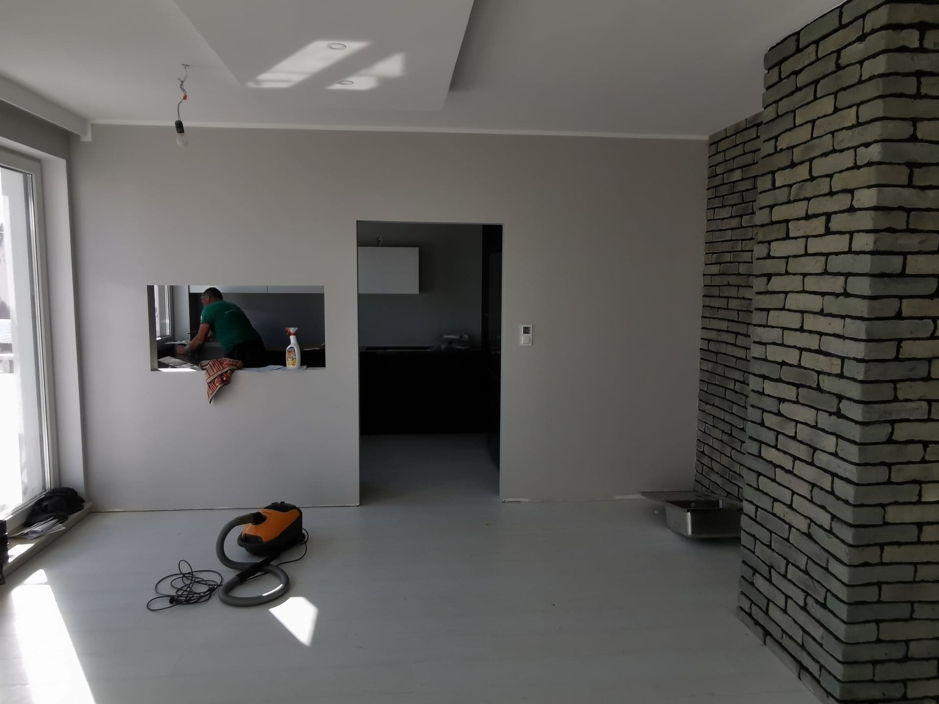 Wnętrze w trakcie prac wykończeniowych: szare ściany, sufit z podwieszanym elementem, dekoracyjna ściana z szarej cegły, pracownik w zielonej koszulce czyści okno, na podłodze odkurzacz.