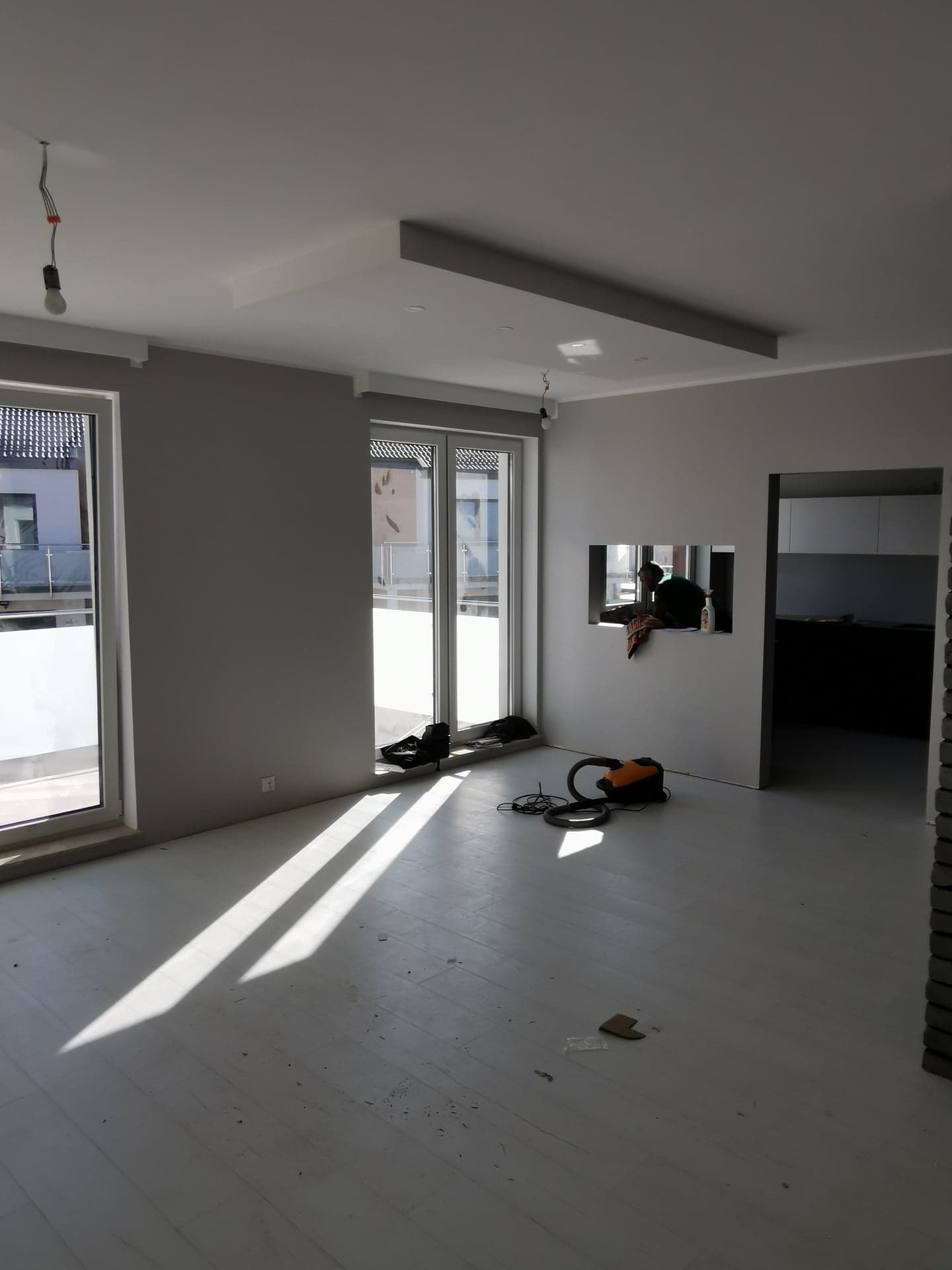 Wnętrze w trakcie remontu: szare ściany, biała podłoga z widocznymi promieniami słońca, otwarte okna balkonowe, odkurzacz i osoba pracująca w otworze ściany.