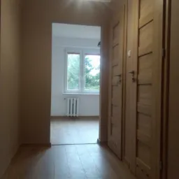 Wnętrze mieszkania po remoncie: widok z korytarza na pokój z drewnianą podłogą, białymi ścianami i grzejnikiem pod oknem, na pierwszym planie drewniane drzwi wewnętrzne z poziomymi panelami.