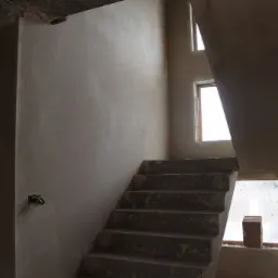 Klatka schodowa w surowym stanie po nałożeniu gładzi, widoczne okna z resztkami zaprawy, wystające kable elektryczne i niedokończone schody.