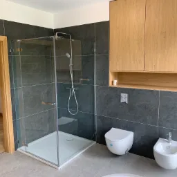 Nowoczesna łazienka z kabiną prysznicową ze szklanymi drzwiami, wiszącą toaletą i bidetem na tle grafitowych płytek i drewnianych szafek.