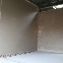 Surowe ściany po nałożeniu tynku, widoczne nierówności przy podłodze, sufit z drewnianych belek i okno dachowe, kabel zwisający ze ściany obok otworu drzwiowego.