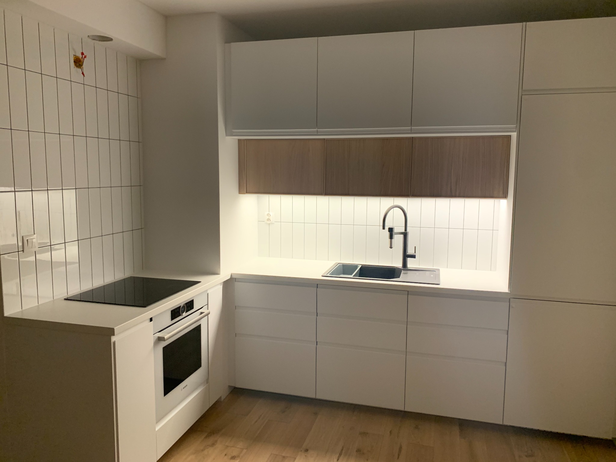 Nowoczesna kuchnia z białymi szafkami, drewnianymi akcentami nad blatem i płytkami w stylu metro, z widoczną instalacją elektryczną na ścianie.