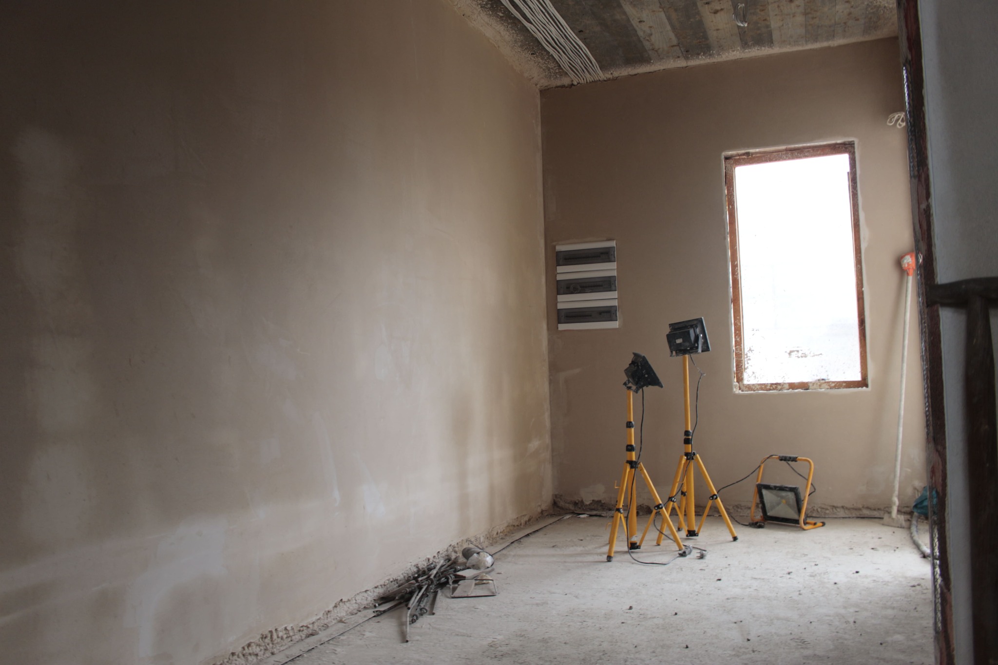 Świeżo otynkowane ściany w jasnym pomieszczeniu z oknem, widoczne przewody pod sufitem, dwie lampy na statywach i rozdzielnia elektryczna na ścianie.