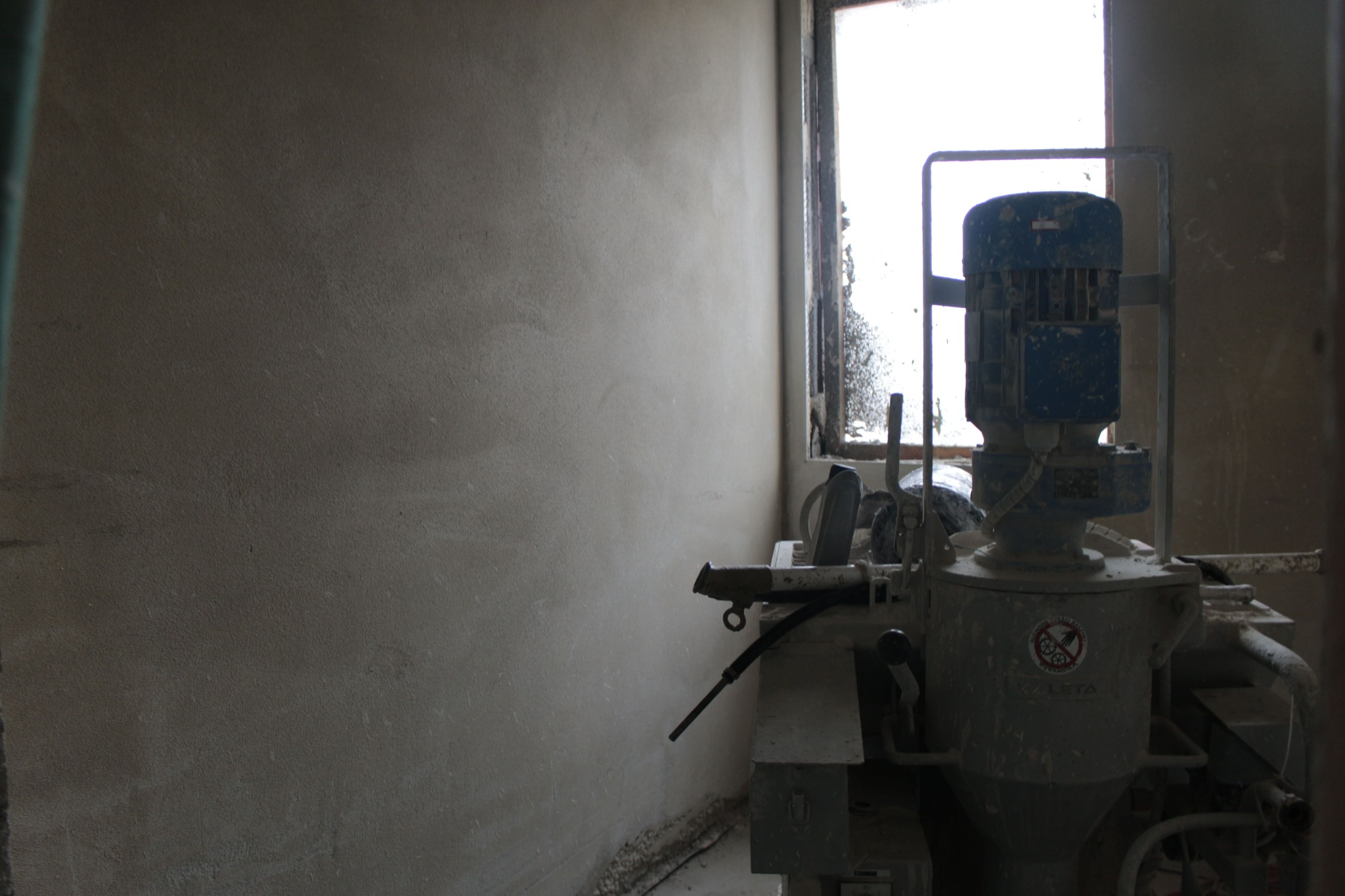 Świeżo nałożony tynk na ścianie w słabo oświetlonym pomieszczeniu, obok stoi brudna maszyna do tynkowania z widocznym silnikiem i naklejką ostrzegawczą.