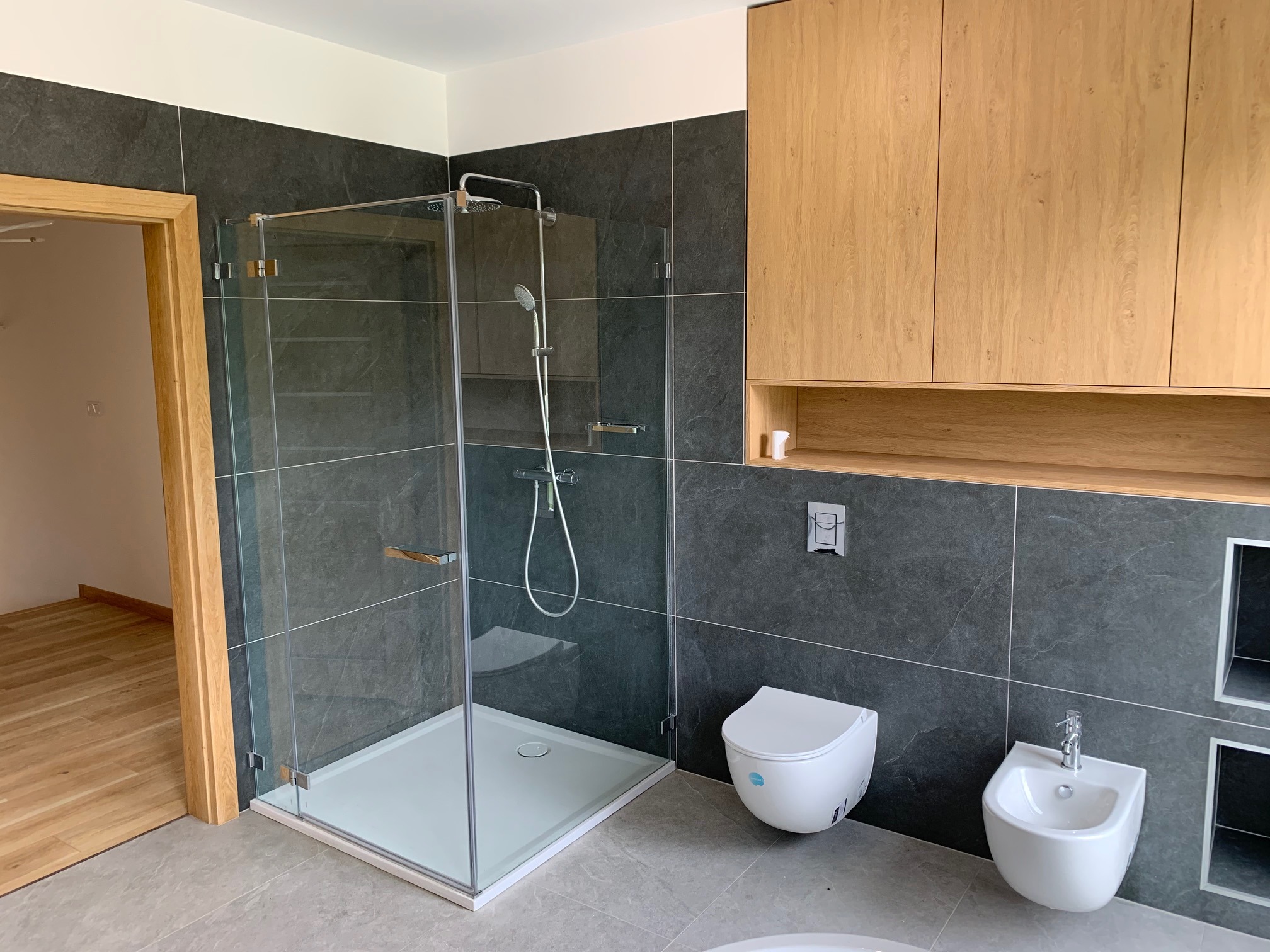 Nowoczesna łazienka z kabiną prysznicową ze szklanymi drzwiami, wiszącą toaletą i bidetem na tle grafitowych płytek i drewnianych szafek.