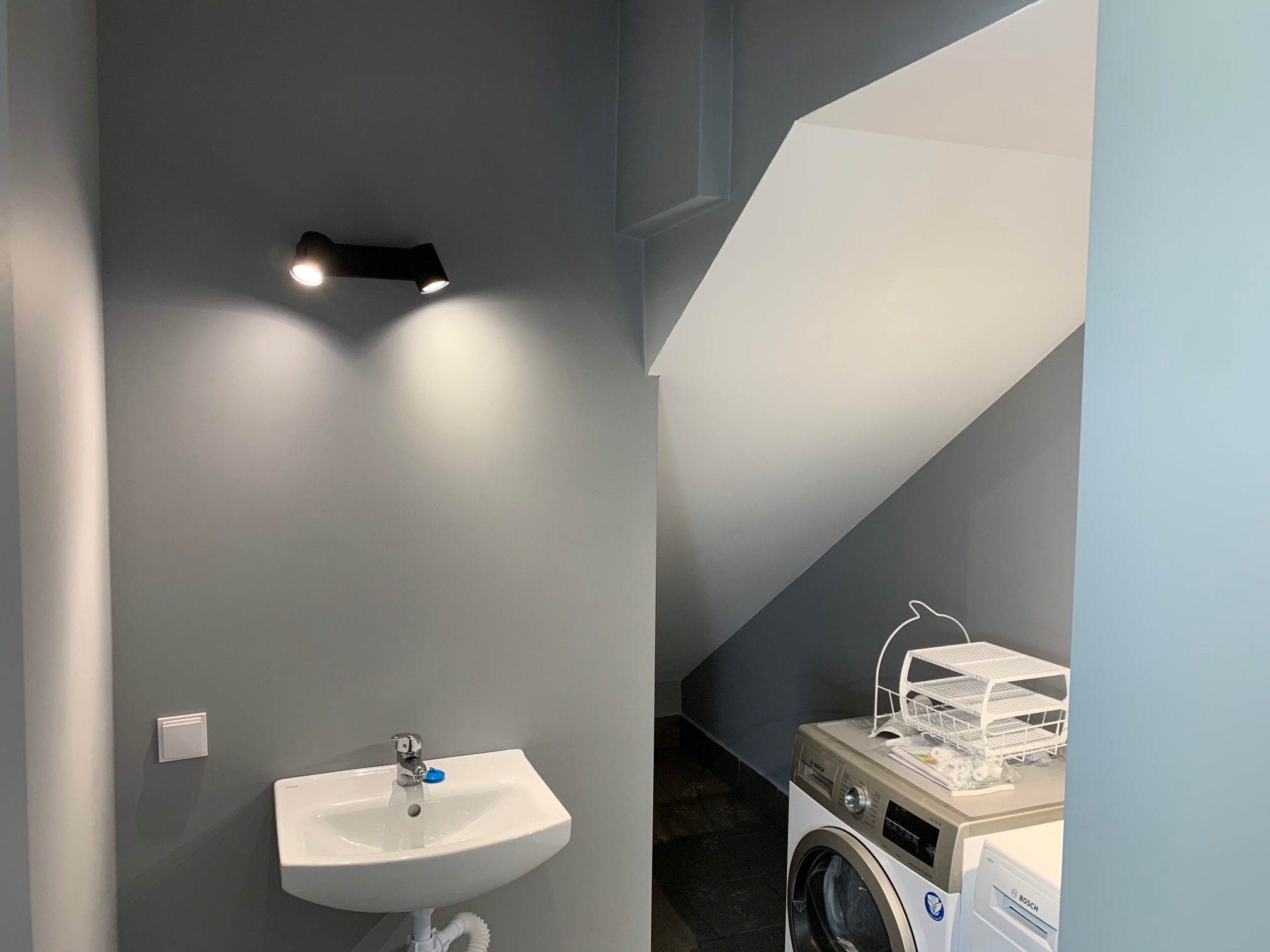 Szara łazienka z umywalką i pralką Bosch pod skosem dachu, oświetlona czarnym reflektorem punktowym.
