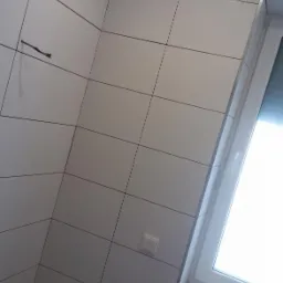 Świeżo położone, białe płytki ceramiczne na ścianach w łazience, widoczne narzędzia na parapecie okiennym i niedokończone prace na suficie.