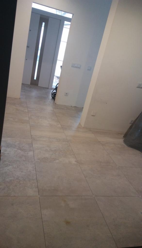 Beżowe płytki podłogowe ułożone w kwadraty, widok z perspektywy podłogi, z widocznym fragmentem drzwi i białymi ścianami w tle.