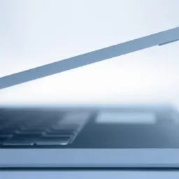 Częściowo otwarty, srebrny laptop w minimalistycznym stylu, z widoczną klawiaturą i touchpadem na tle jasnego, rozproszonego światła.