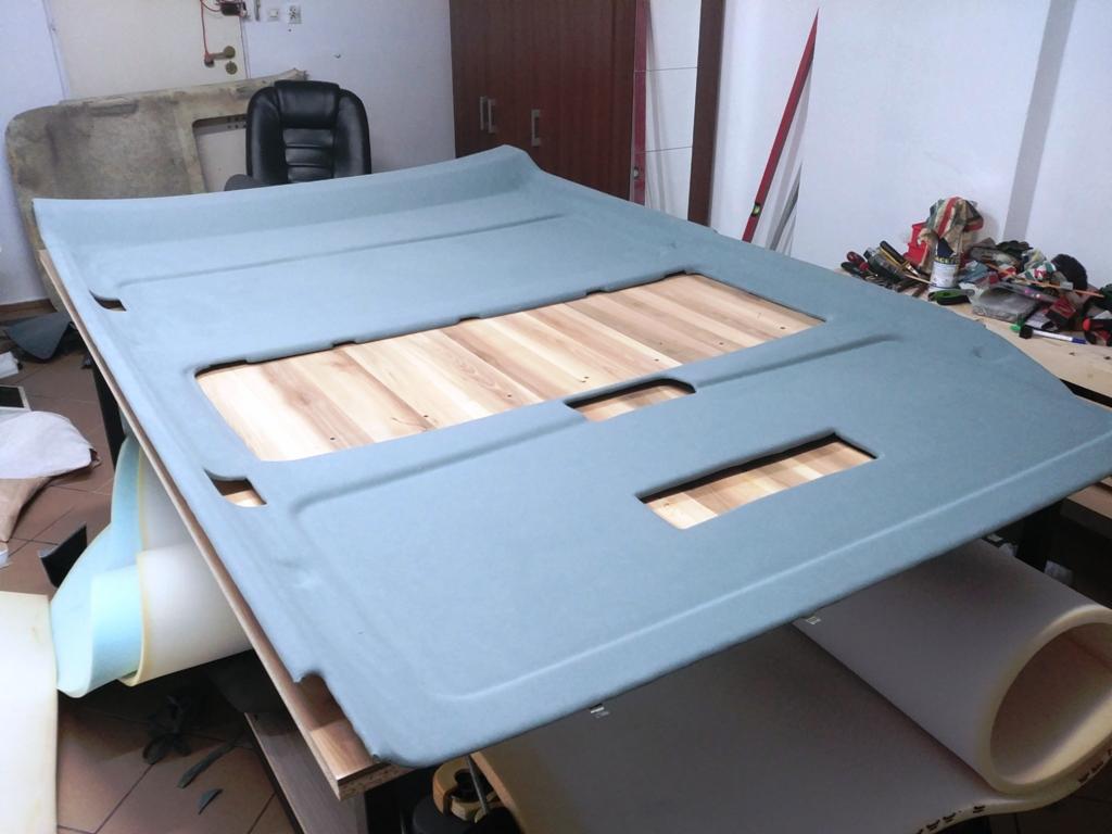 Szary podsufitka samochodowa w trakcie prac renowacyjnych, leżąca na drewnianym stole warsztatowym, widoczne materiały tapicerskie i narzędzia w tle.