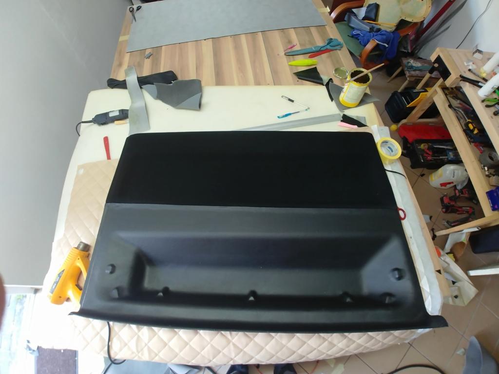 Panel samochodowy z czarną tapicerką, częściowo obity, leżący na stole warsztatowym z narzędziami i materiałami.