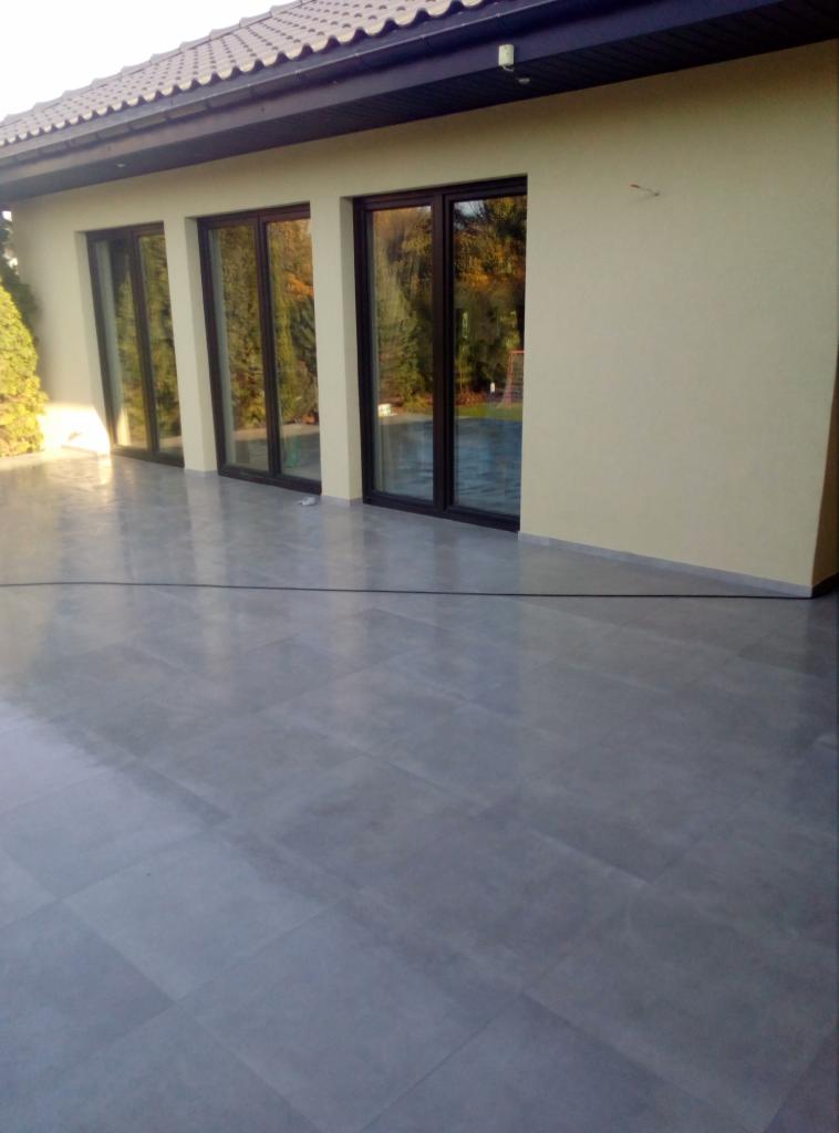 Szare płytki gresowe na tarasie przylegającym do domu z brązowymi oknami tarasowymi.