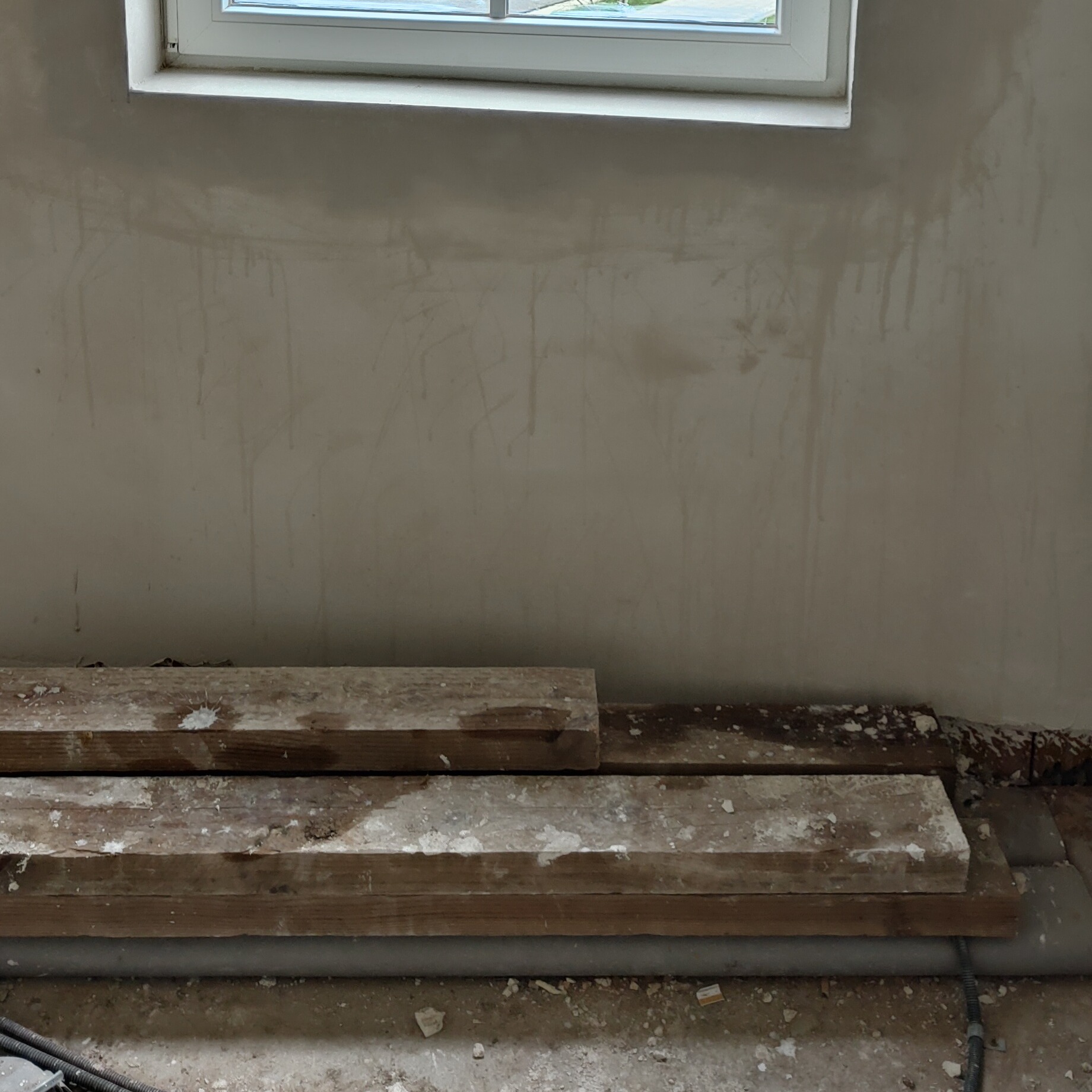 Świeżo nałożony tynk na ścianie pod oknem, z widocznymi śladami po nałożeniu. Na podłodze leżą drewniane deski i fragmenty gruzu.