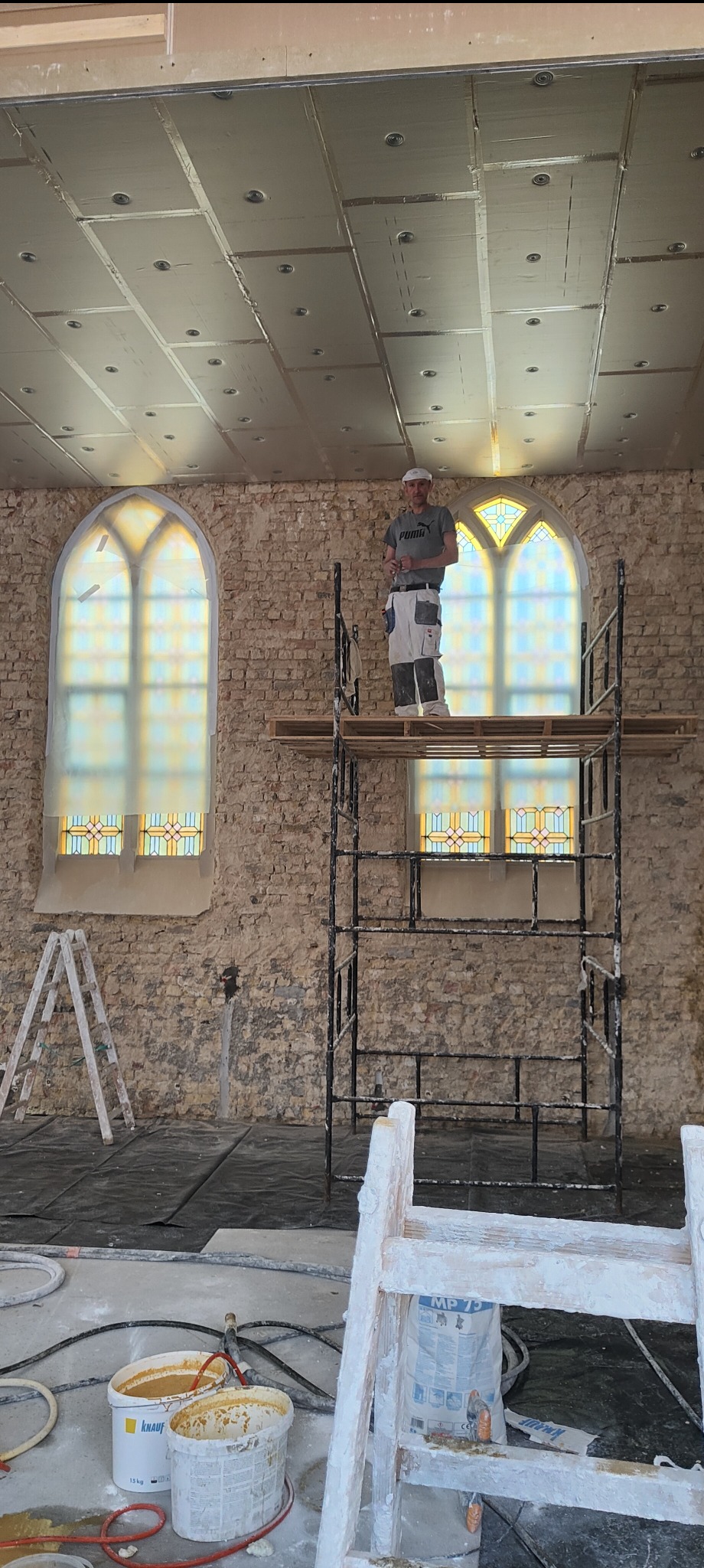 Pracownik na rusztowaniu w trakcie prac wykończeniowych w pomieszczeniu z ceglanymi ścianami i witrażowymi oknami. Widoczne przygotowane materiały i narzędzia.