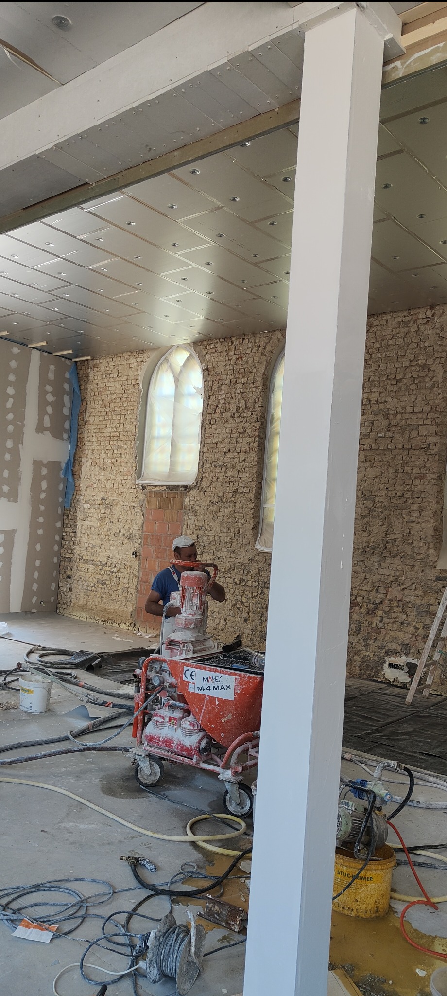 Pracownik w trakcie aplikacji tynku maszynowego na ścianę z cegły, używając pomarańczowej maszyny. Widoczne okna zabezpieczone folią i sufit z izolacją.