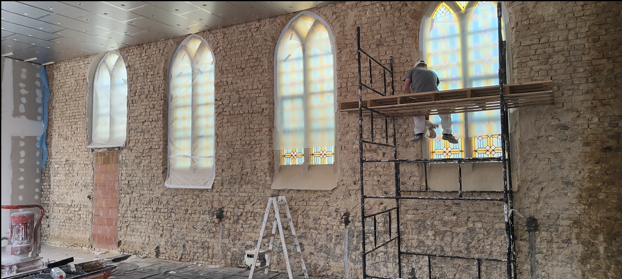 Prace tynkarskie w Nasiadkach: murarz na rusztowaniu, przygotowuje ścianę z cegły z gotyckimi oknami do tynkowania maszynowego. Widoczne narzędzia i zabezpieczenia okien.