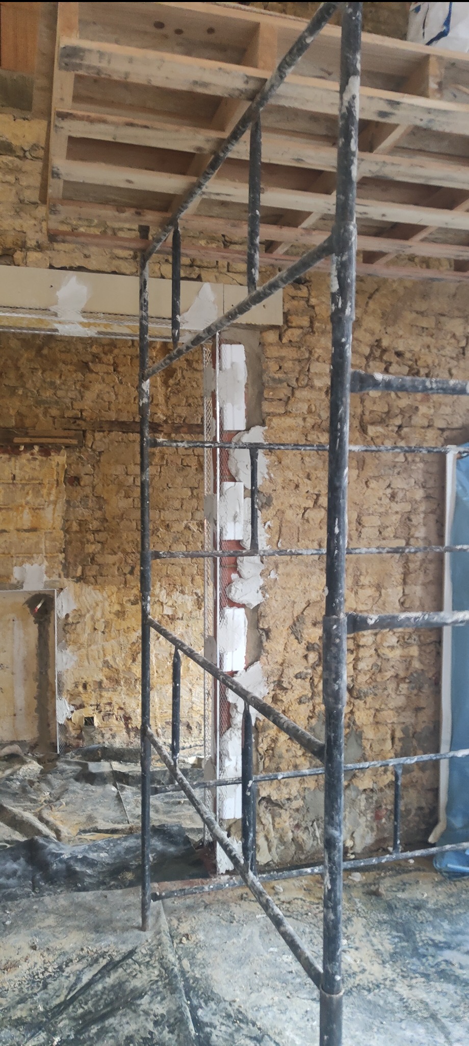 Przygotowanie ściany z cegły do tynkowania: fragment ściany z nałożoną siatką i zaprawą, widoczne rusztowanie. Prace murarskie w trakcie realizacji.