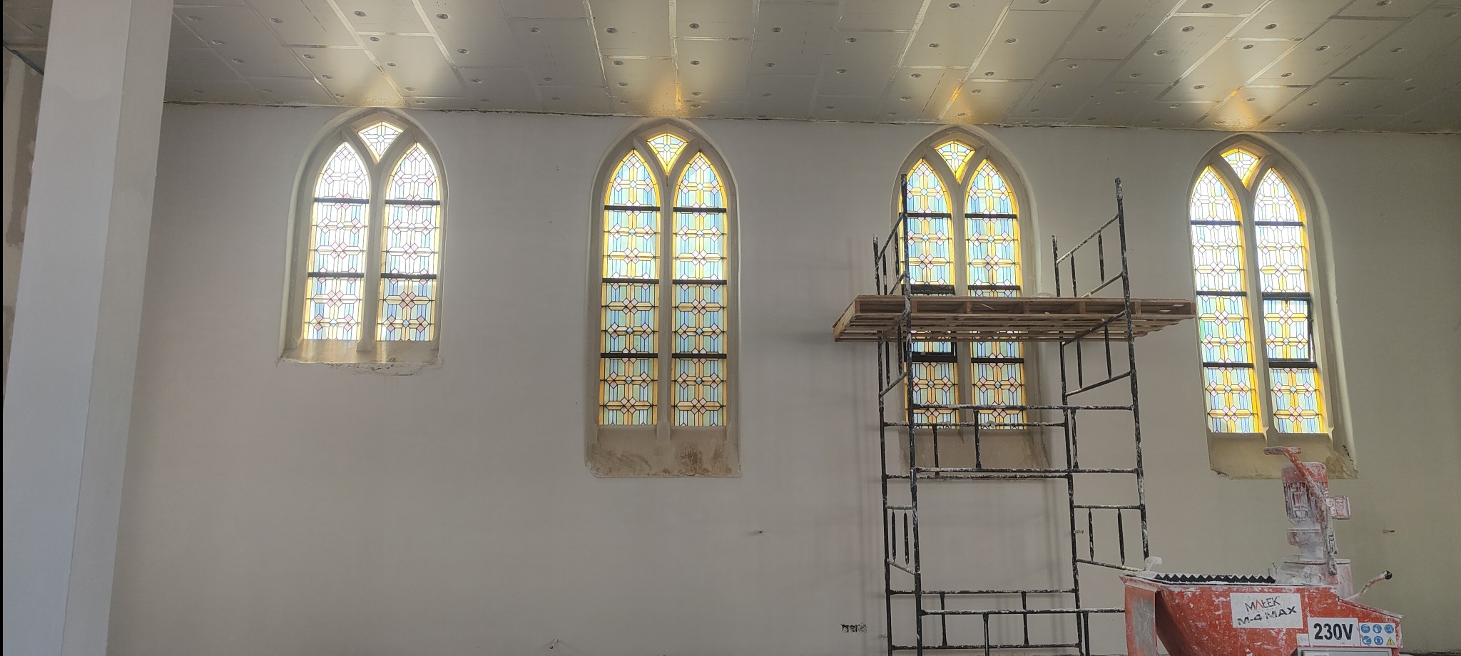 Wnętrze kościoła w trakcie prac. Widoczne witraże w oknach, rusztowanie i maszyna do tynkowania. Ściany przygotowane do nałożenia tynku.