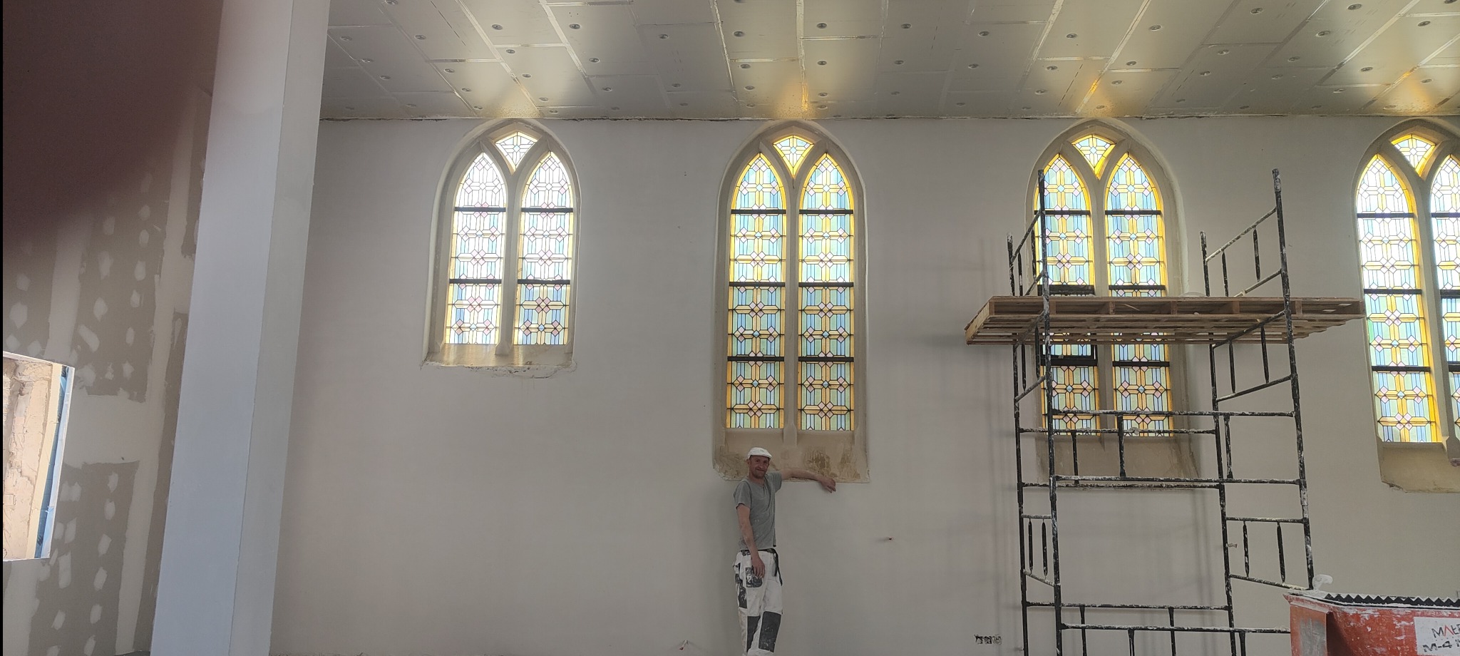 Wnętrze kościoła w trakcie prac tynkarskich. Widoczne witraże, rusztowanie i pracownik w ubraniu roboczym. Ściany przygotowane do dalszych prac wykończeniowych.