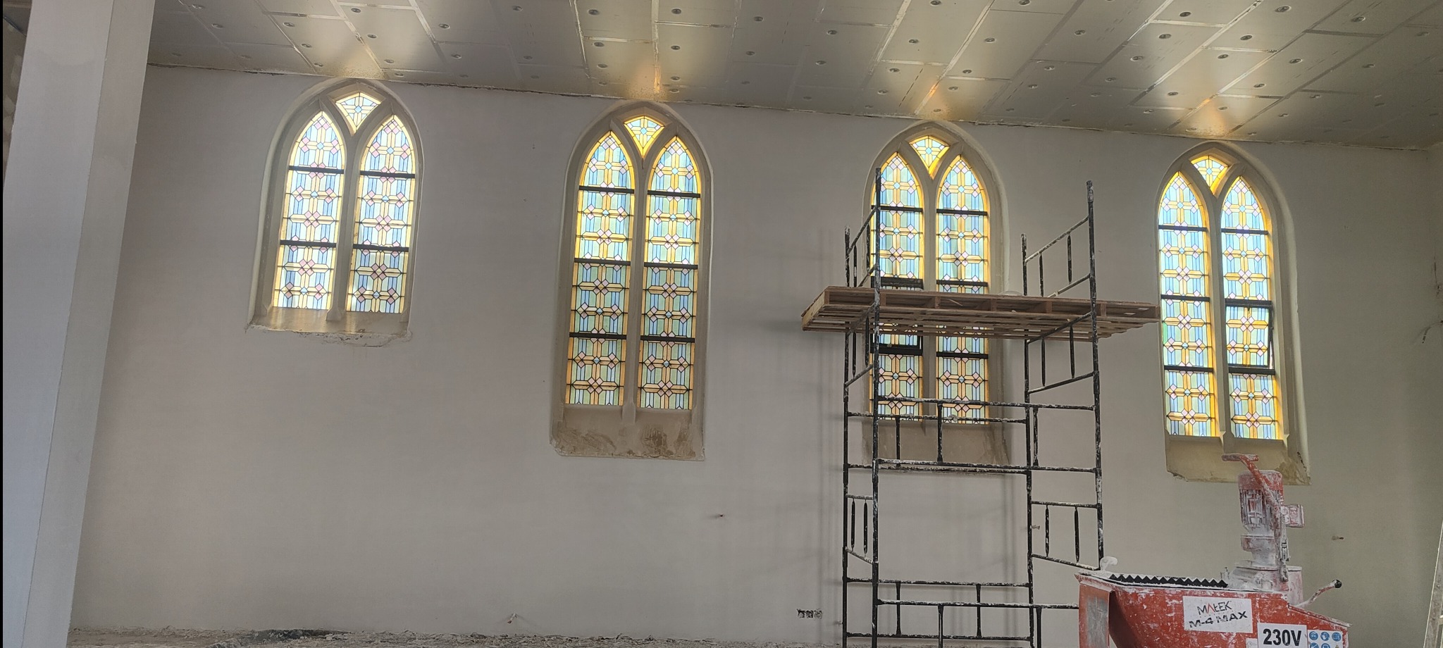Wnętrze kościoła w trakcie prac tynkarskich: rusztowanie przed witrażowym oknem, maszyna do tynkowania, ściany po nałożeniu tynku. Widoczne ślady prac.