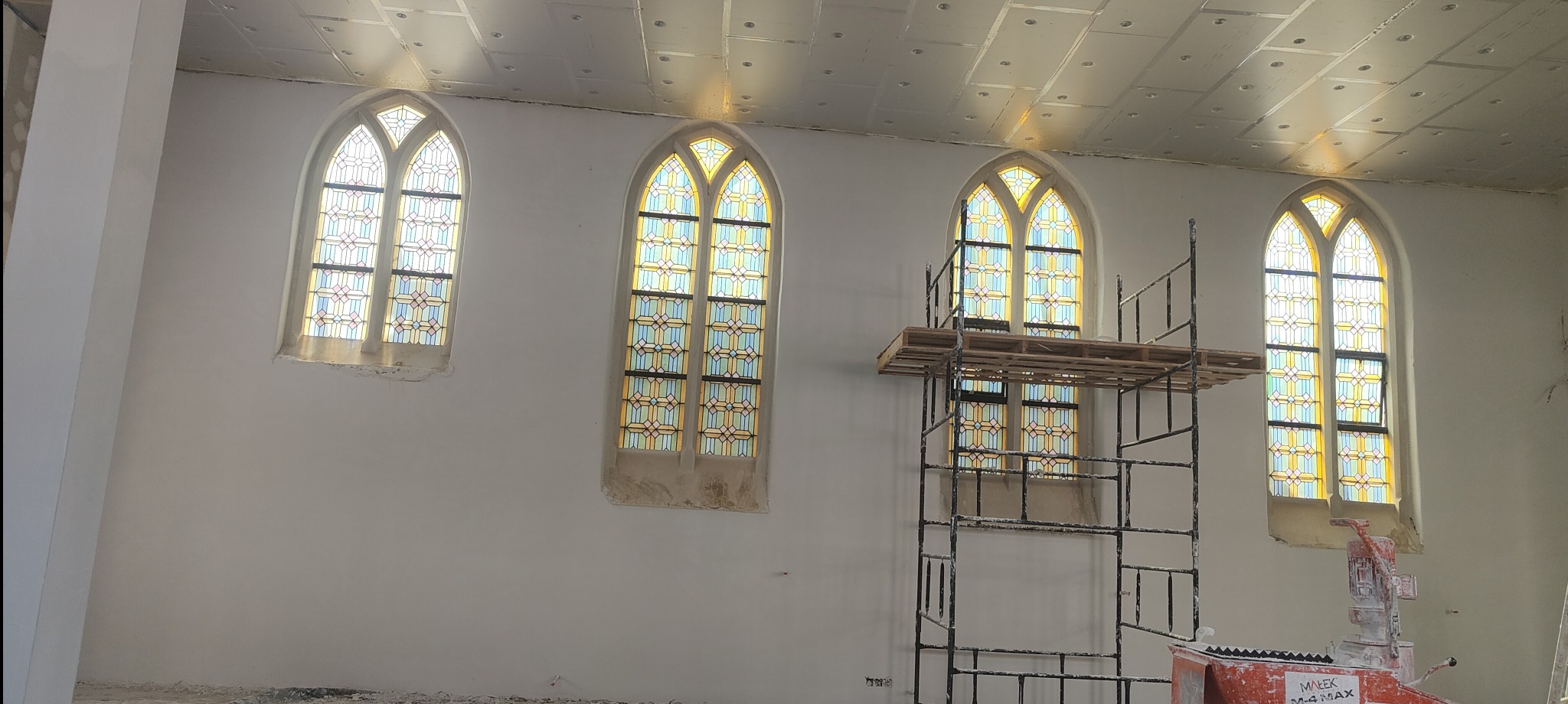 Wnętrze kościoła w trakcie tynkowania maszynowego: ściany po tynkowaniu, rusztowanie, witraże w oknach, maszyna do tynkowania.