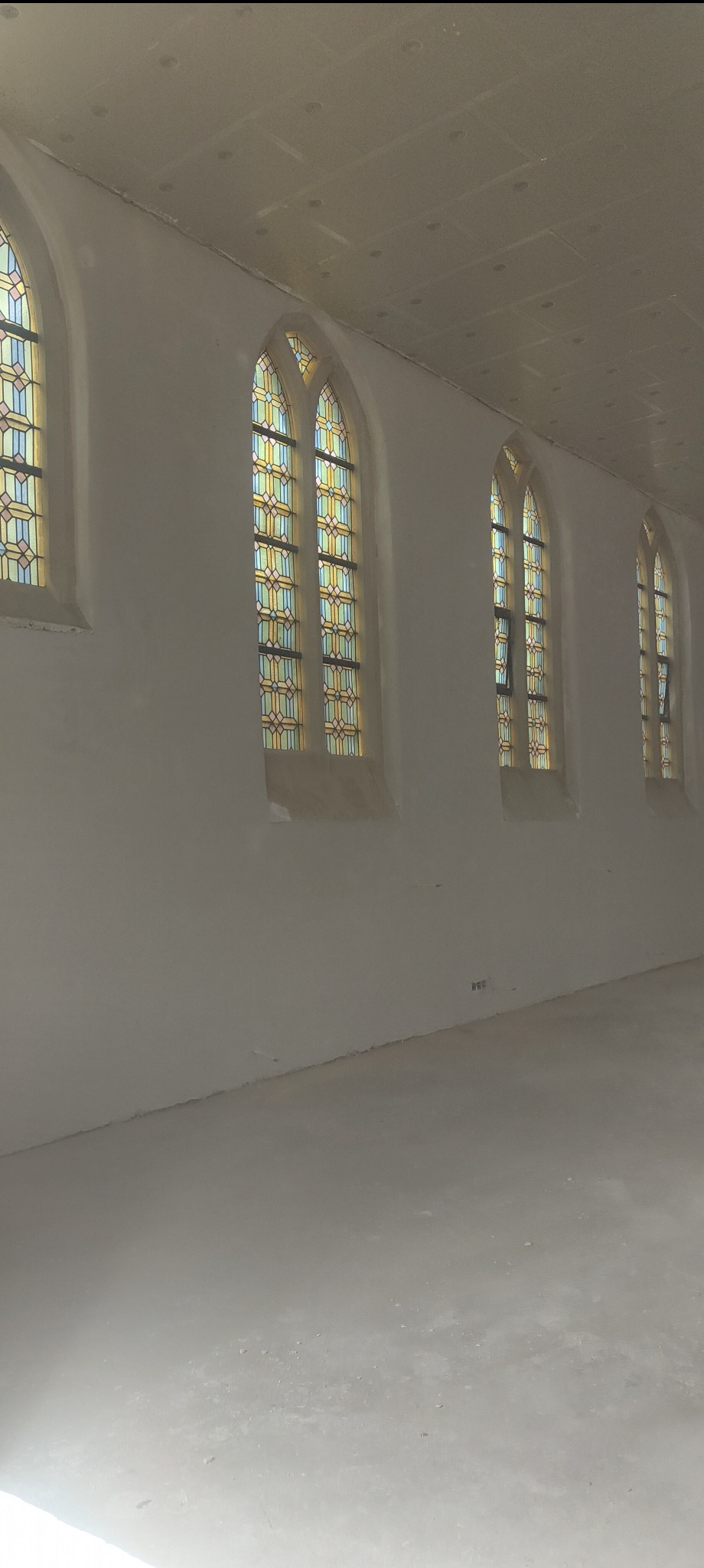Wnętrze po tynkowaniu maszynowym: gładkie, jasne ściany z wysokimi oknami witrażowymi w gotyckim stylu. Widoczne ślady po pracach wykończeniowych na podłodze.