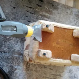 Czyszczenie tapicerowanego pufy od spodu za pomocą odkurzacza piorącego; widoczne drewniane nóżki i fragment dywanu w tle.