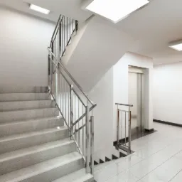 Wnętrze klatki schodowej w budynku biurowym z widocznymi schodami z szarego lastryko, metalową balustradą, białymi ścianami, płytkami podłogowymi i windą.