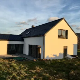 Nowoczesny dom jednorodzinny z jasną elewacją i ciemnym, metalowym dachem, widziany z perspektywy trawnika w promieniach zachodzącego słońca; w tle zalesione wzgórza i samochód dostawczy z reklamą...