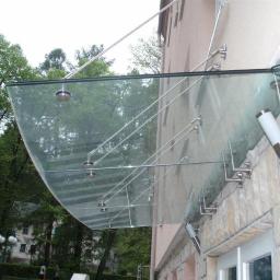 Jarom - Szklany daszek nad wejściem do budynku, mocowany na stalowych wspornikach i linkach, widok z dołu, z oświetleniem zewnętrznym.