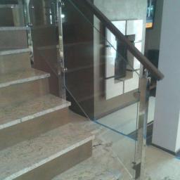 Jarom - Szklana balustrada z elementami ze stali nierdzewnej i drewnianą poręczą zamontowana przy schodach z jasnego kamienia. Widoczne ślady po remoncie na podłodze.