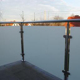 Jarom - Balkon z nowoczesną balustradą ze szkła matowego i stali nierdzewnej, widok z zewnątrz, szara płytka na podłodze, w tle domy z czerwonymi dachami i drzewa.