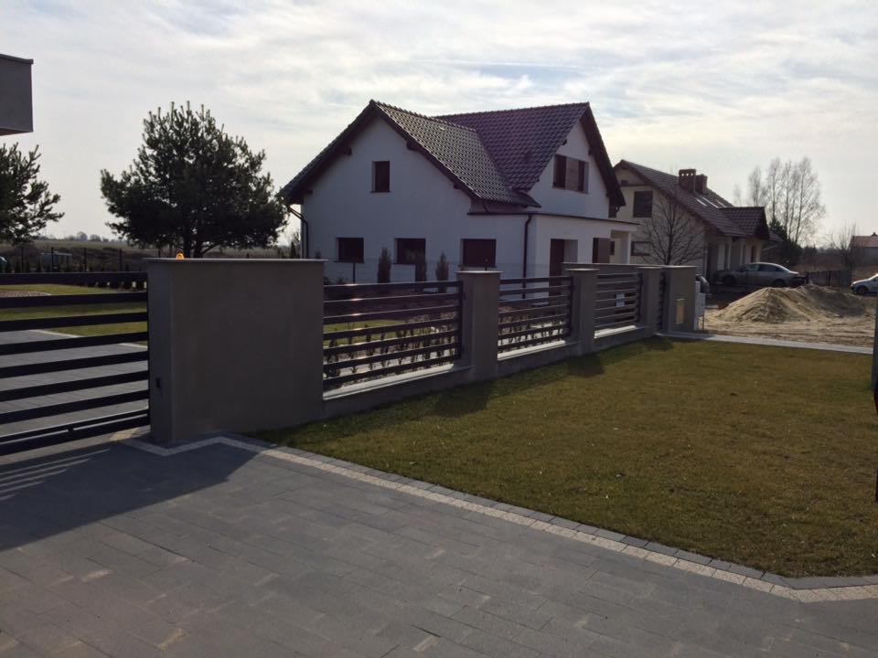 Nowoczesne ogrodzenie posesji z poziomych, ciemnych profili metalowych osadzonych między betonowymi słupkami, na tle domu jednorodzinnego z ciemną dachówką i trawnika.