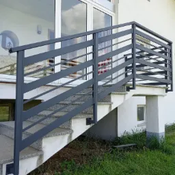 Zewnętrzne schody z szarego granitu z nowoczesną, grafitową balustradą z poziomych profili, prowadzące do przeszklonych drzwi z widocznym odbiciem flagi USA w szybie