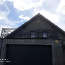 Nowoczesny dom z płaskim dachem garażu, na którym znajduje się balkon z metalową balustradą z poziomych belek, widok z dołu na tle błękitnego nieba z chmurami.