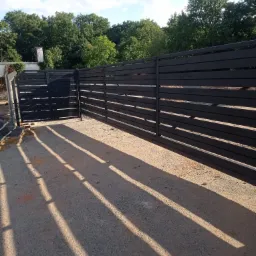 Nowoczesne, poziome ogrodzenie metalowe w kolorze antracytowym z przesuwną bramą wjazdową, rzucające długie cienie na szutrową nawierzchnię podjazdu, w tle zieleń drzew.