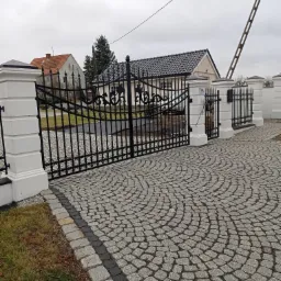 Czarna, ozdobna brama wjazdowa z metalu z białymi, murowanymi słupkami, prowadząca na posesję z brukowanym podjazdem ułożonym w wachlarz.