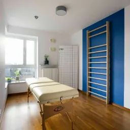 Jasny gabinet fizjoterapii z beżowym stołem do masażu, drabinką gimnastyczną na niebieskiej ścianie i oknem z widokiem na miasto.