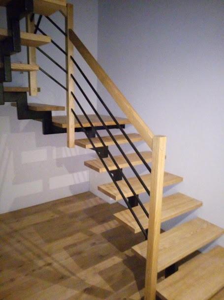 Schody drewniane z balustradą z czarnych, cienkich prętów, zamocowanych do drewnianej poręczy i stopni, oświetlone światłem padającym z boku.