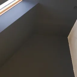 Pomieszczenie na poddaszu z oknem dachowym, pomalowane na ciemny szary kolor, widoczny fragment ściany w stanie surowym przed malowaniem.