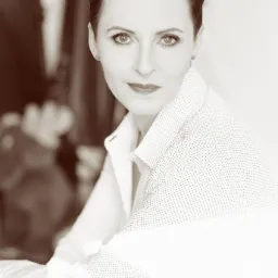 Portret kobiety z krótkimi włosami w białej koszuli w kropki, z delikatnym uśmiechem, w sepiowej tonacji.