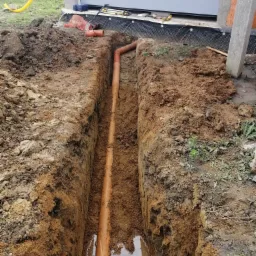 Wąski wykop w ziemi z pomarańczową rurą kanalizacyjną na dnie, widoczne narzędzia i żółty przewód gazowy w tle przy budynku.