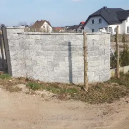 Metalowa brama wjazdowa z ozdobnym elementem, przylegająca do murku z szarej kostki, dalej ogrodzenie z siatki na betonowej podmurówce, w tle domy jednorodzinne.