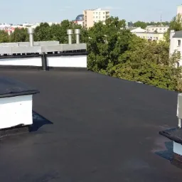 Czarny, płaski dach pokryty papą termozgrzewalną z białymi kominami wentylacyjnymi i metalowymi zakończeniami, widok na panoramę miasta w tle.