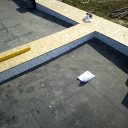 Układanie izolacji z płyt OSB i styropianu na dachu pokrytym papą, widoczna poziomica i opakowania materiałów.